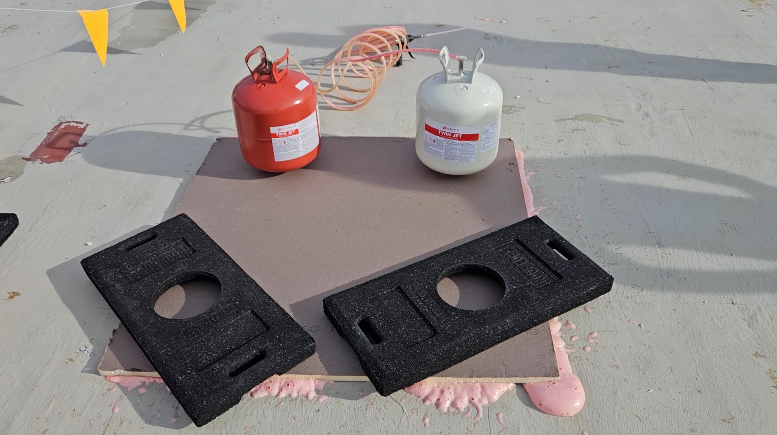 Elevate Twin Jet two-component urethane adhesive canisters and weighted rubber pavers set on a polyisocyanurate board, with pink adhesive squeezed out around the board edges